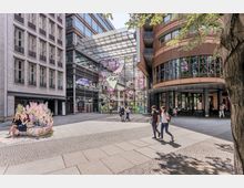 Potsdamer Platz in Berlin mit modernen Gebäuden, einer großen Glasfassade und dekorativen Blumenillustrationen. Menschen spazieren und sitzen auf einer bunten Kunstinstallation vor dem Haupteingang unter einem schattigen Baum.
