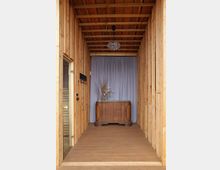 Schmaler Innenraum mit Holzwänden und Holzdecke, am Ende steht eine antike Holzkommode mit dekorativen Trockenblumen darauf. Im Hintergrund hängt ein grauer Vorhang, und von der Decke hängt ein Kronleuchter.