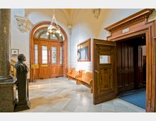 Eingangsbereich eines historischen Gebäudes mit Holztüren und verzierten Glasfenstern, Marmorboden und einer Büste auf einem Sockel. Rechts befindet sich eine offene Tür mit der Aufschrift „Loge N“ sowie Holzsitzbänke an der Wand.
