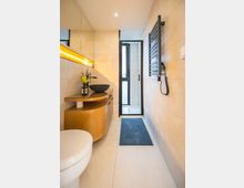 Modernes Badezimmer mit hellen Fliesen, einem hölzernen Waschtisch mit Aufsatzwaschbecken und leuchtender LED-Spiegelbeleuchtung. Der Raum verfügt über einen schwarzen Handtuchheizkörper, eine gläserne Duschtür und einen blauen Teppichläufer.