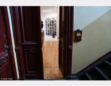 Blick durch eine geöffnete, dunkelrote Holztür in ein Zimmer mit hellem Holzboden. Im Hintergrund befindet sich ein Regal mit vielen Schuhen, und ein Hund sitzt auf einem hellen Teppich.