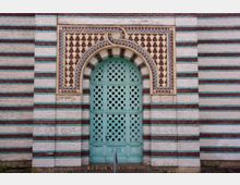 Fassade mit orientalischem Stil: Eine grünlich-türkise Tür mit filigranen, sternförmigen Mustern und einem Spitzbogen, umrahmt von dekorativen Keramikfliesen in Gelb-, Blau- und Brauntönen. Die Wand besteht aus horizontalen Streifen aus weißem, braunem und grünem Backstein.