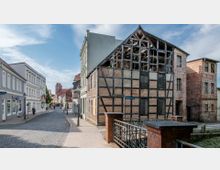 Straßenszene in einer Altstadt mit Kopfsteinpflaster, Fachwerkhäusern und modernen Gebäuden. Im Vordergrund ein teils zerfallenes Fachwerkhaus, rechts ein Geländer aus Metall und Ziegelsteinen, im Hintergrund ein Kirchturm.