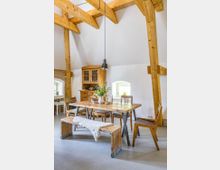 Esszimmer in einem rustikalen Stil mit Holzbalkendecke, einem Esstisch aus massivem Holz, Stühlen und einer Holzbank mit einer Decke. Im Hintergrund ein Holzschrank und kleine Fenster mit weißen Rahmen, durch die Tageslicht einfällt.