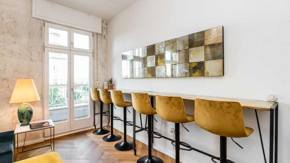 Heller Innenraum mit langem Hochtisch entlang einer weißen Wand, davor mehrere Barhocker mit senfgelben Polstersitzen und einer großen rechteckigen Wandkunst aus spiegelnden Kacheln. Links führen hohe Glastüren auf einen Balkon; der Raum hat Fischgrätparkett, eine Stehlampe und eine teils unverputzte Wandfläche.