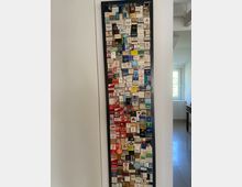 Schwarzer Wandrahmen mit einer Sammlung von Streichholzschachteln in verschiedenen Größen, Farben und Designs. Der Rahmen befindet sich in einem hellen Innenraum mit weißer Wand und sichtbarem Fenster im Hintergrund.