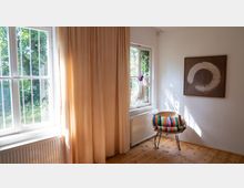 Helles Zimmer mit Holzfußboden, großen Fenstern mit weißen Rahmen und beigefarbenen Vorhängen. An der Wand hängt ein Bild mit einem abstrakten weißen Kreis, daneben steht ein Stuhl mit buntem Streifenkissen.