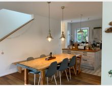 Essbereich und offene Küche in einem modernen Wohnhaus. Ein großer Holztisch mit sechs Stühlen steht auf einem Parkettboden, im Hintergrund eine helle Küche mit einer Insel, Holzarbeitsflächen und Hängelampen, sowie ein Fenster mit Blick ins Grüne.