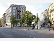 Straßenkreuzung in einer Stadt mit historischen Gebäuden im Gründerzeitstil, flankiert von Straßenbäumen. Im Vordergrund sind Gehwege, eine Bushaltestelle sowie Fußgänger und parkende Autos sichtbar.