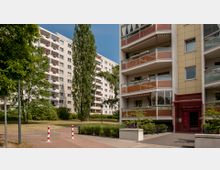 Mehrstöckige Wohngebäude mit Balkonen, umgeben von Grünflächen, Bäumen und Gehwegen. Im Vordergrund ist ein markierter Weg mit rot-weißen Pollern zu sehen, der zu einem Eingangsbereich mit roter Tür führt.