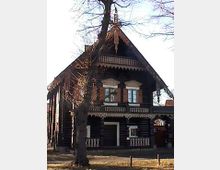 Holzhaus im traditionellen Stil mit steilem Satteldach und dekorativen Verzierungen an der Dachkante. Vor dem Haus steht ein Baum, und die Umgebung wirkt ländlich mit einem offenen, grasbewachsenen Bereich.