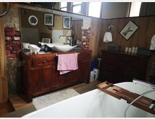 Rustikales Badezimmer mit Holz- und Backsteinelementen. Im Vordergrund steht eine freistehende Badewanne, dahinter eine Holzkommode mit Waschbecken und Spiegel, umgeben von dekorativen gerahmten Bildern an der Wand.