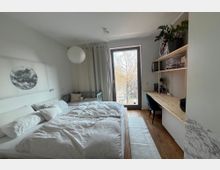 Schlafzimmer mit schlichtem, modernem Design: Im Vordergrund ein Doppelbett mit weißer Bettwäsche, daneben ein Kinderbett. Rechts ein Schreibtisch aus Holz mit Regalen, Grünpflanzen und Arbeitsmaterialien. Im Hintergrund ein großes Fenster mit Blick auf Bäume und eine Straße.