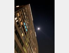 Ein hohes, modernes Hochhaus mit beleuchteten Fenstern ragt schräg in den Nachthimmel empor, mit dem Vollmond und dunklem, wolkenlosem Hintergrund.