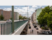 Städtische Straßenszene mit einer erhöhten Brücke links und einer darunter verlaufenden, schmaleren Straße. Rechts sind mehrstöckige Wohnhäuser mit Balkonen und Bäumen zu sehen, links stehen größere Wohngebäude mit einheitlicher Architektur.