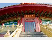 Eingangsbereich eines traditionellen chinesischen Gebäudes mit rotem Dach, kunstvoll verzierten Dachstreben und grünen Fenstern. Eine Treppe mit gelbem Geländer und dekorativen Steinskulpturen führt zum Eingang, über dem eine rote Tafel mit goldenen Schriftzeichen hängt.