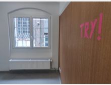 Innenansicht eines Raumes mit weiß gestrichenen Wänden, einem rechteckigen Fenster mit Sprossen, das nach draußen auf eine Ziegelwand blickt, und einer darunter angebrachten Heizkörper. Rechts ist eine Holzplatte zu sehen, an der in pinkem Klebeband das Wort "TRY!" angebracht ist.