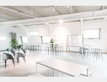 Heller, moderner Seminarraum mit weiß gestrichenen Wänden und Sichtbetondecke, ausgestattet mit Stühlen, hohen Tischen, Barhockern, einem Whiteboard auf Rollen und Zimmerpflanzen. Große, schräg eingesetzte Fenster lassen viel Tageslicht herein.