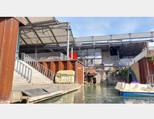 Eine Szenerie in einem ehemaligen Industriegebäude mit modernen Überdachungen aus Metall und Glas. Im Vordergrund sind eine Treppe, ein Steg mit Wasserzugang und Tretboote auf einem Wasserbecken zu sehen, während Teile einer alten Backsteinfassade und rostige Stahlelemente die Kulisse bilden.