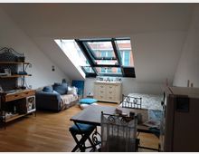 Ein Wohnzimmer unter einer Dachschräge mit einem großen Fenster, das auf ein gegenüberliegendes Backsteingebäude blickt. Der Raum enthält eine Couch, ein Metallbett, einen Esstisch mit Stühlen, einen kleinen Schrank und ein Regal; der Boden besteht aus hellbraunem Holz.