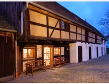 Historisches Fachwerkgebäude bei Dämmerung, mit beleuchteten Fenstern und Holztüren, umgeben von einem gepflasterten Hof. Der obere Teil ist aus Holz und Lehm, der untere aus weiß gestrichenem Mauerwerk.