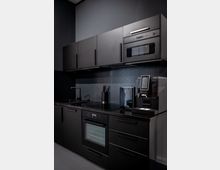 Moderne, dunkelgraue Einbauküche mit matten Schrankfronten, schwarzer Arbeitsplatte und schlichter Rückwand. Sichtbar sind Spüle, Backofen und Mikrowelle sowie eine Kaffeemaschine auf der rechten Seite der Arbeitsfläche.