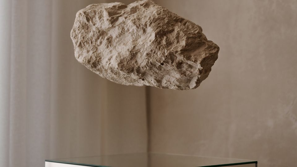 Innenraum mit minimalistischer Präsentation: Ein schwebender, rauer Steinblock hängt über einem würfelförmigen Sockel aus spiegelndem Glas. Im Hintergrund sind eine beige Wand und ein halbtransparenter Vorhang zu sehen.