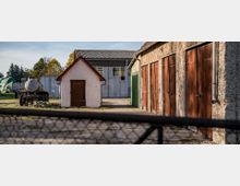 Ein ländlicher Hof mit mehreren Gebäuden, darunter eine kleine Hütte mit Holztür, eine Reihe von Garagen mit Holztoren und ein modernes, metallverkleidetes Gebäude im Hintergrund. Im Hof steht ein alter Anhänger mit einem Tank, und verschiedene landwirtschaftliche Geräte sind teils abgedeckt.