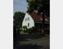 Ein weißes Wohnhaus mit rotem Satteldach und mehreren Fenstern, umgeben von einem Holzzaun und dichter Vegetation. Im Vordergrund ein gepflasterter Weg, daneben Bäume, die teilweise Schatten spenden.