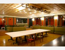 Innenraum eines Veranstaltungsraums mit Holzboden, einer großen weißen Tischplatte auf kunstvoll verzierten Tischbeinen und mehreren Stühlen. Die Wände sind in Grün- und Orangetönen gehalten, mit integrierter Bar, Stufenelementen und Wandlampen; die Decke ist mit einem geometrischen Muster dekoriert, mit mehreren Scheinwerfern installiert.