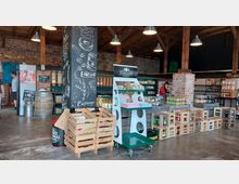 Ein rustikaler Hofladen mit Backsteinwänden und Holzbalkendecke, in dem regionale Produkte wie Säfte, Marmeladen und Getränke angeboten werden. Im Vordergrund stehen Holzkisten und ein Verkaufsständer, während im Hintergrund Regale mit weiteren Waren zu sehen sind.