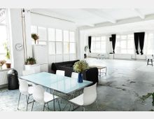 Offener, heller Raum in einem industriellen Loftstil mit weißen Wänden und großen Fenstern, die von schwarzen Vorhängen eingerahmt sind. Im Vordergrund steht ein Esstisch mit Glasplatte und weißen Stühlen, dahinter eine schwarze Couch sowie Pflanzen und Dekorationen, während der hintere Bereich weitläufig und minimalistisch gehalten ist.