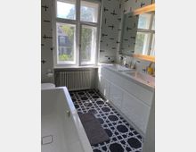Modernes Badezimmer mit großen Fenstern, die viel Tageslicht hereinlassen. Das Zimmer verfügt über eine freistehende Badewanne, einen Waschtisch mit Spiegel und weißen Schränken sowie schwarz-weiß geflieste Wände und einen geometrisch gemusterten Boden.