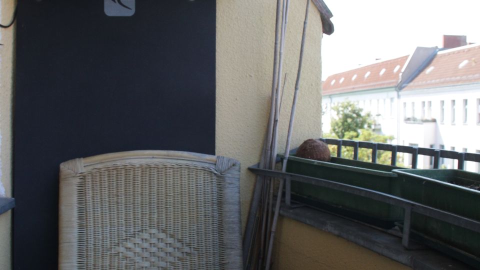 Small apartment balcony with yellow stucco walls, a rustic stick railing above, and a black door panel. A wicker armchair sits beside a weathered wooden table with a glass ashtray, and a railing with planter boxes overlooks nearby buildings.