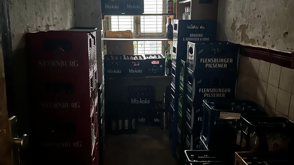 Schmaler Getränkelagerraum mit hohen Stapeln aus Getränkekisten entlang der Wände und Regalbrettern mit Flaschen vor einem großen, vergitterten Fenster. Abgenutzte Wände und ein gewölbter Deckenbereich betonen den kellerartigen Charakter des Raums.