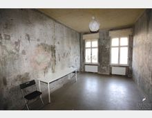 Karger, leerer Innenraum mit abgenutzten, fleckigen Putzwänden und blankem Boden, spärlich möbliert mit einem langen Tisch und Klappstühlen. An der Stirnseite befinden sich zwei hohe Fenster mit Heizkörpern darunter und eine einfache Deckenleuchte.