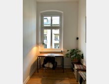 Kleines, schlichtes Arbeitszimmer mit Holzboden und einem schmalen Fenster, das Tageslicht hereinlässt. Ein einfacher Holzschreibtisch mit Lampe, Laptop und Schreibutensilien steht vor dem Fenster; daneben befindet sich eine grüne Zimmerpflanze und rechts eine gepolsterte Sitzbank.