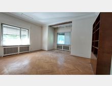 Ein leerer Raum mit hellem Fischgrätparkettboden, großen Fenstern mit Jalousien und Heizkörpern darunter. Rechts steht ein eingebautes Holzregal, an der Decke sind Schäden durch herausbrechendes Putzmaterial sichtbar.