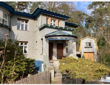 Zweistöckiges Wohnhaus im Stil der frühen Moderne, mit grauer Putzfassade, einer überdachten, bunt verglasten Eingangsterrasse und blauen Dachkanten. Im Vorgarten stehen Sträucher, und rechts ist ein separates Garagengebäude mit gelber Dacheinfassung zu sehen, umgeben von Bäumen.