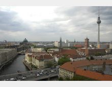Stadtansicht von Berlin mit historischen Gebäuden und modernen Highlights, darunter der Berliner Fernsehturm, der Berliner Dom mit seiner Kuppel und das Rote Rathaus. Im Vordergrund fließt die Spree, umgeben von roten Ziegeldächern und Brücken, während am Horizont Wohn- und Bürogebäude zu sehen sind.