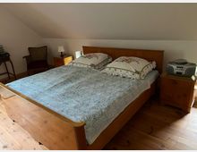 Schlafzimmer mit einem holzfarbenen Doppelbett, dekoriert mit floral gemusterten Kissenbezügen und einer gemusterten Tagesdecke. Links befinden sich ein antiker Stuhl und ein kleiner Tisch mit Büchern, rechts ein Nachttisch mit einem Radio-CD-Player. Der Raum hat Holzboden und weiße Wände.