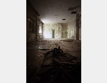 Ein verlassener Raum mit beschädigter Decke und Wänden, deren Putz großflächig abblättert. Der Boden ist mit Trümmern und Schutt bedeckt, während am Ende des Raumes ein schmaler Türdurchgang sichtbar ist.