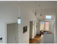 Flur mit weißen Wänden und Holzboden, deckenhängenden Lampen, mehreren Türen an der linken Wand und einem Fenster mit weißen Vorhängen am Ende des Raumes. Eine schräge Decke mit Dachfenster lässt zusätzliches Licht herein, und ein kleines Tischchen mit Gegenständen steht am Fenster.