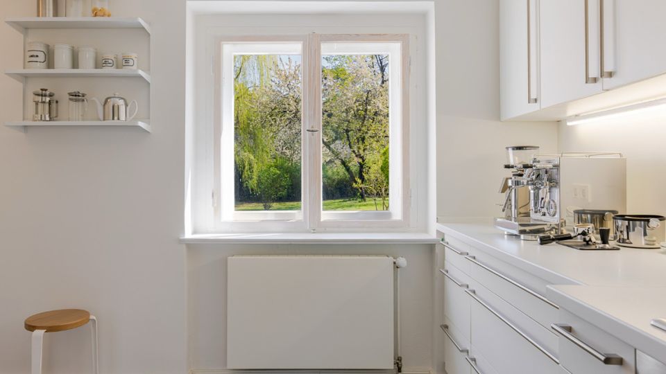 Helle, minimalistische Küche mit weißen Schränken, grauem Fliesenboden und einer langen Arbeitsplatte mit Kaffeemaschine. Ein großes Fenster über dem Heizkörper blickt auf einen grünen Garten; links sind offene Wandregale mit Gläsern und Dosen.