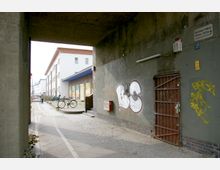 Unterführung mit grauer Wand, die mit Graffiti besprüht ist, daneben eine braune Metalltür. Rechts stehen geparkte Fahrräder vor einem flachen Gebäude mit weißer Fassade und blauen Vordächern. Im Hintergrund sind weitere moderne Wohngebäude und eine Baustellenabsperrung sichtbar.