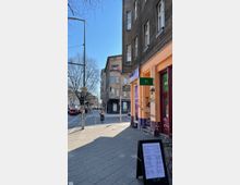 Straßenszene in einem urbanen Viertel mit mehrstöckigen Altbauten. Im Vordergrund ein Gehweg mit einem aufgestellten Menüschild vor einem orangefarbenen Gebäude, daneben eine Straßenecke mit Radfahrern, Bäumen und Verkehrsschildern.