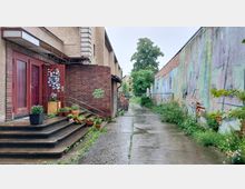 Eine Seitengasse mit einer gepflasterten Fläche, links ein Eingang mit roten Türen, einer Backsteinwand und Treppen dekoriert mit Blumentöpfen. Rechts eine bemalte Mauer mit verwitterten Stellen, im Hintergrund Bäume und ein Zaun.