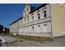 Altes Wohngebäude mit hellgrauer Fassade, zahlreichen Fenstern mit teils heruntergelassenen Rollläden und schlichtem Stuckdekor. Vor dem Gebäude befinden sich ein unbefestigter Gehweg, eine kleine Grünfläche und eine Bushaltestelle.