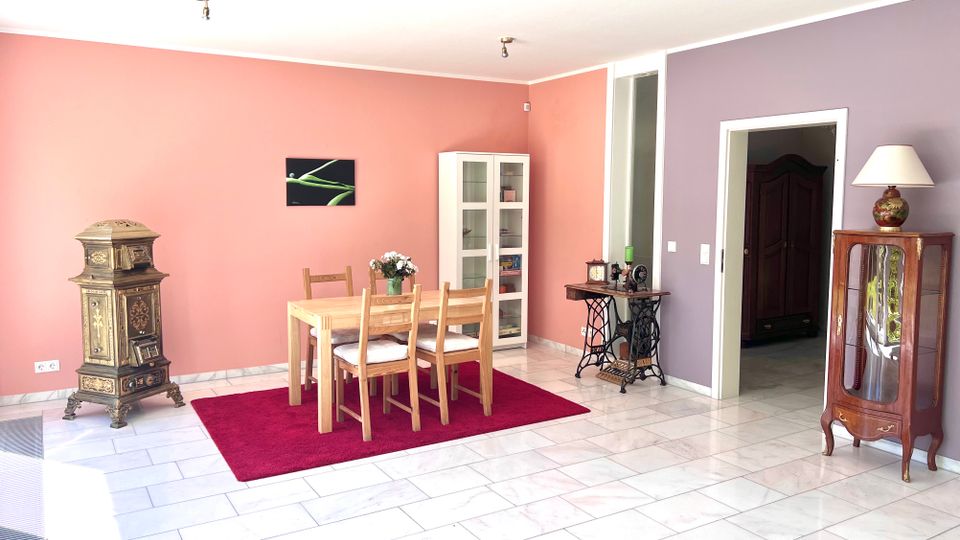 Heller Essbereich mit rosafarbener und lilafarbener Wand, einem Holzesstisch mit vier Stühlen auf rotem Teppich und hellem Fliesenboden. Im Raum stehen ein antiker Ofen, eine Vitrine sowie ein kleiner Tisch mit Nähmaschinenuntergestell, und rechts führt eine Tür in einen Nebenraum.