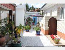 Ein gepflegter Innenhof mit hellem Kies, eingerahmt von weiß verputzten Gebäuden und Klinkersteinfassaden. Der Hof ist mit zahlreichen Pflanzen in farbigen Töpfen dekoriert, darunter ein Sonnenschirm, ein Holztisch mit Stühlen und eine blaue Tür im Hintergrund.